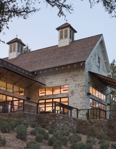 Modern rustic building at dusk with illuminated interiors and exterior sconces, featuring a stone base, large windows, and weathered wood siding.