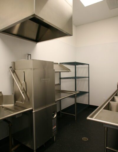 A clean and empty commercial kitchen with stainless steel equipment, including sinks, a dishwasher, and storage racks.