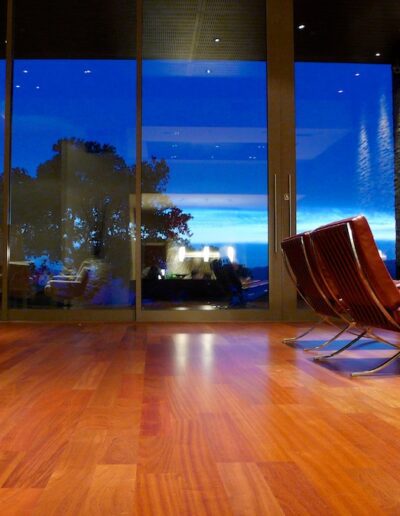 Modern living room with wooden flooring, large windows, and a single red chair at twilight.