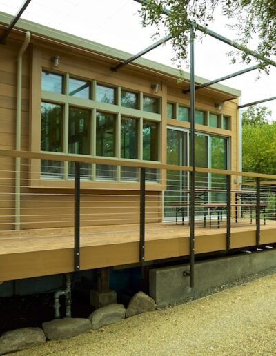 A modern wooden building with a large glass facade and a deck featuring cable railings, surrounded by greenery.