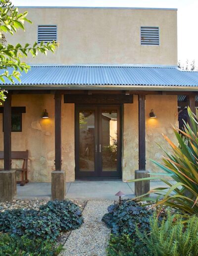 A single-story stucco building with a metal roof surrounded by xeriscaped gardens and a tree.