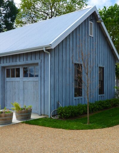 A rustic blue barn with a metal roof adjacent to a gravel path and green landscaping.