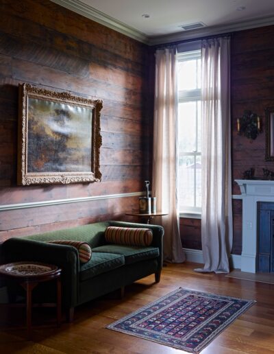 A cozy corner of a room featuring a dark wood-paneled wall, a vintage-style olive green couch with a striped cushion, an oriental rug, light-draping curtains, and classic artwork.